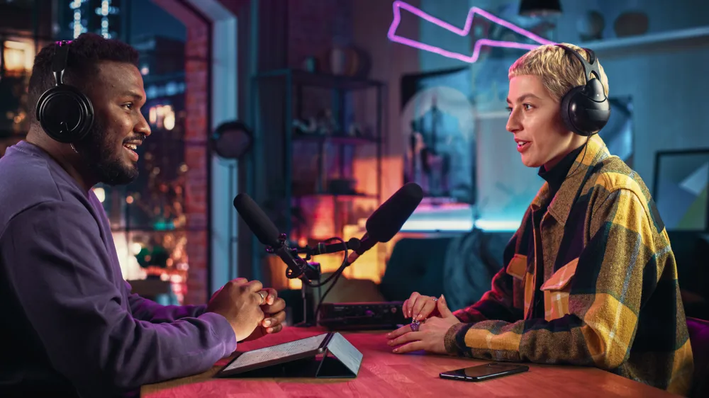 ALT Text: Two Young Stylish Radio Show Hosts Record Fresh Podcast Episode in Home Loft Studio Apartment. Attractive Energetic Co-hosts Discuss Important Topics Live on Air in an Evening Show.