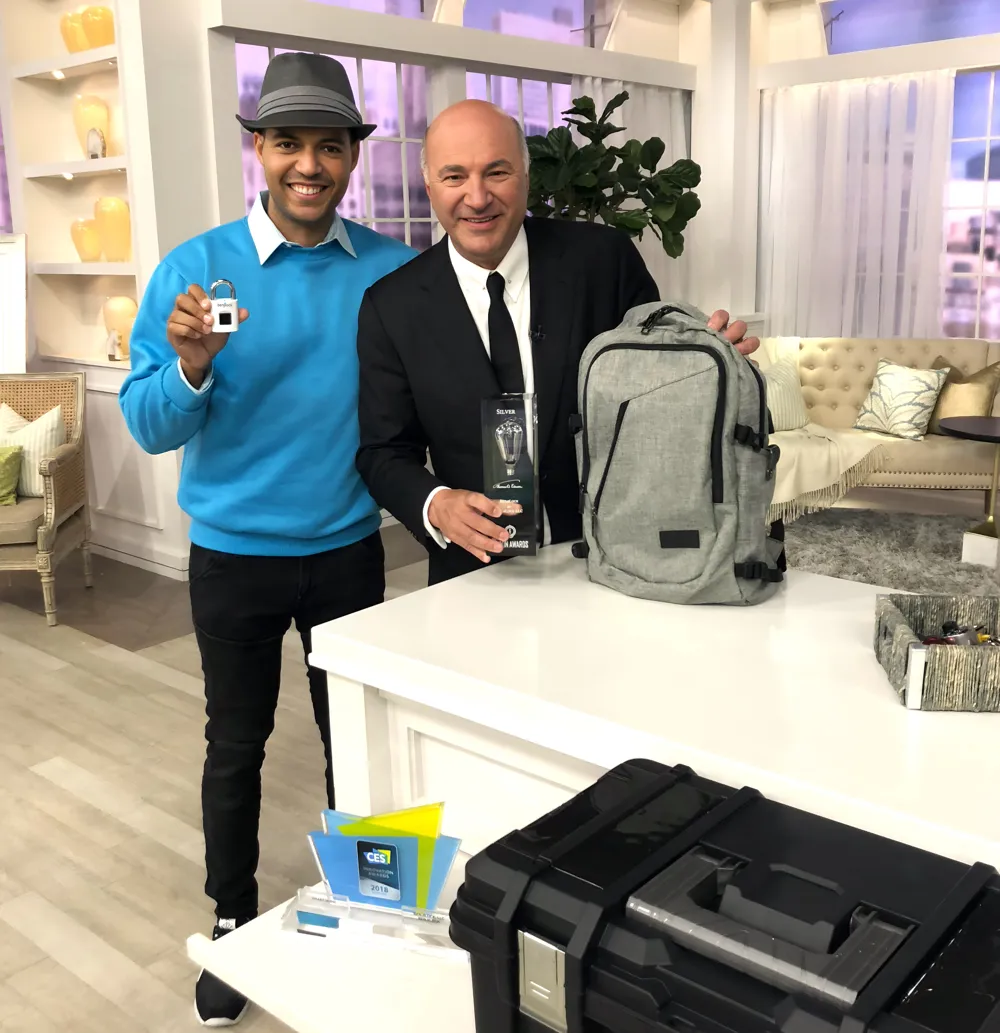 Robbie Cabral, the inventor of BenjiLock, and businessman Kevin O'Leary (from Shark Tank) stand next to a display table. Cabral, wearing a blue sweater and a hat, holds up a small BenjiLock. O'Leary, in a suit, holds a small trophy-like award next to a grey backpack. A black, rugged case is in the foreground.